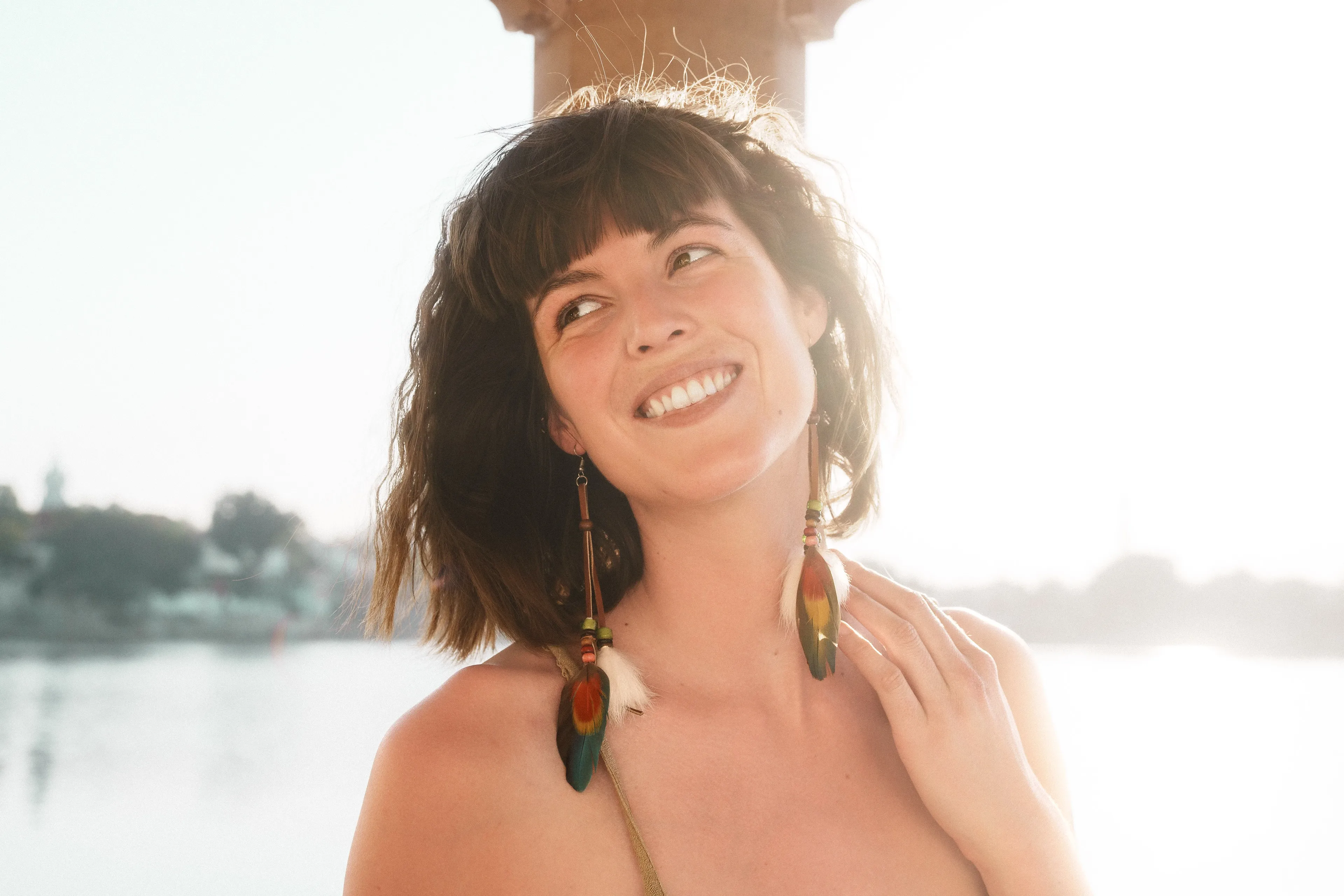 Backlit portrait of a woman with feather earrings, smiling by the water