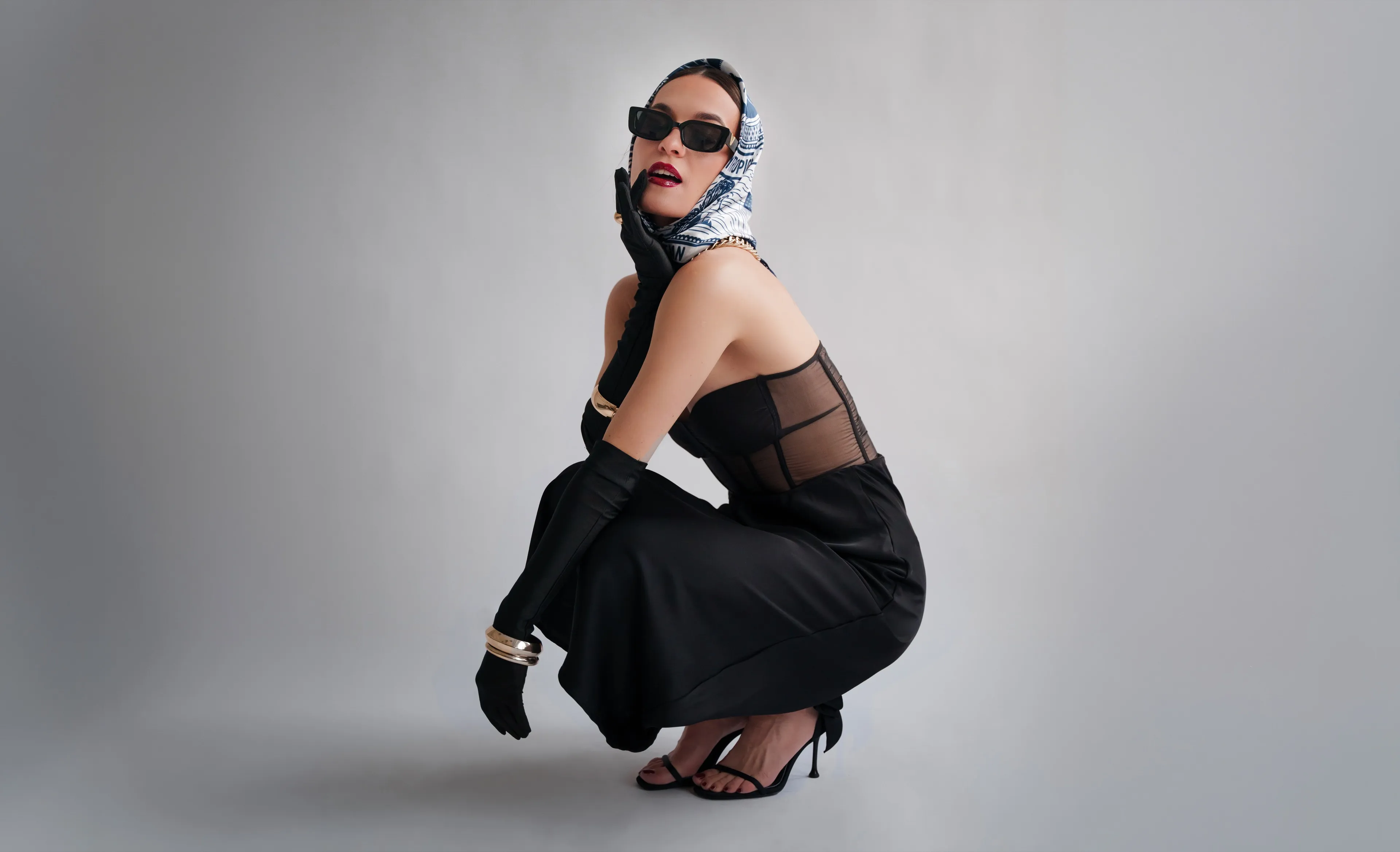 Editorial portrait — a model crouching in a black dress, headscarf and sunglasses against a grey studio wall