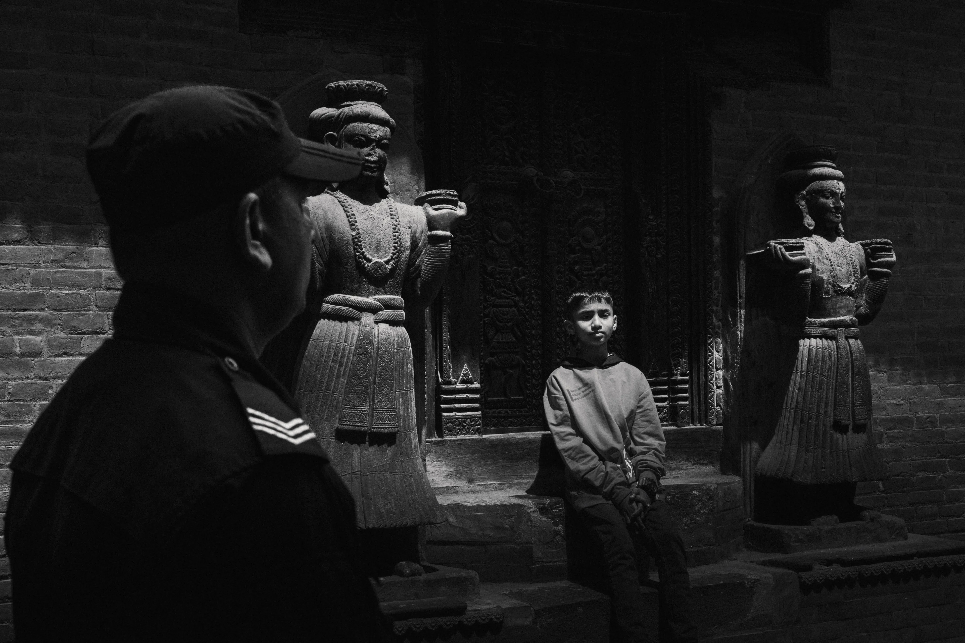 Black and white street photograph — a uniformed man in the foreground, a boy seated between carved temple guardians